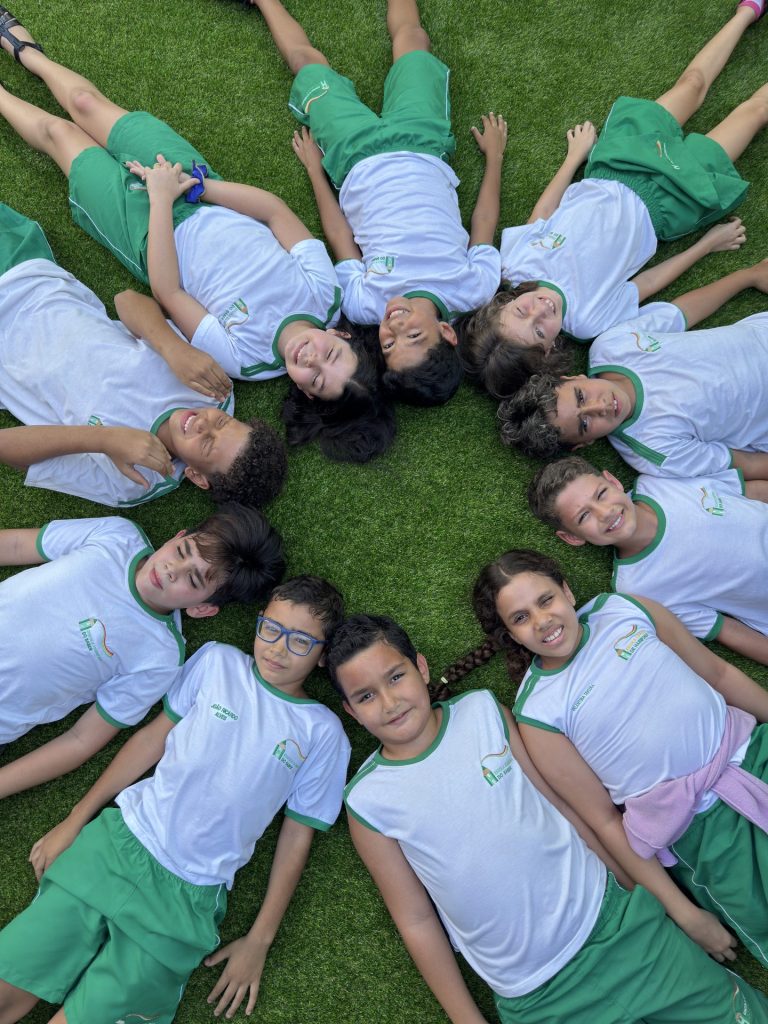 Alunos de uniforme deitados em círculo no gramado da escola